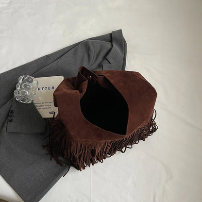 Dark brown suede shoulder bag with fringe, on gray blazer, clear glass, and a book.