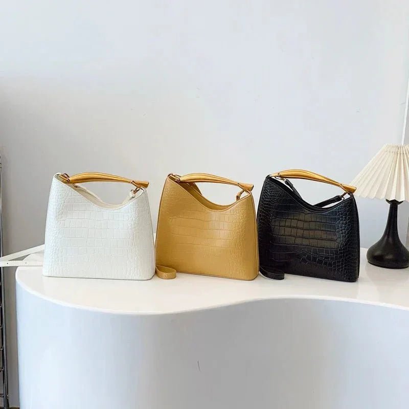 Three crocodile pattern handbags in white, camel, and black with top handles on a white table.