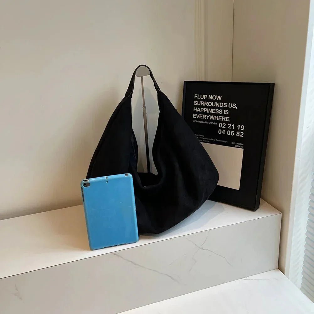 Large black suede hobo bag with zipper and blue tablet on a white shelf
