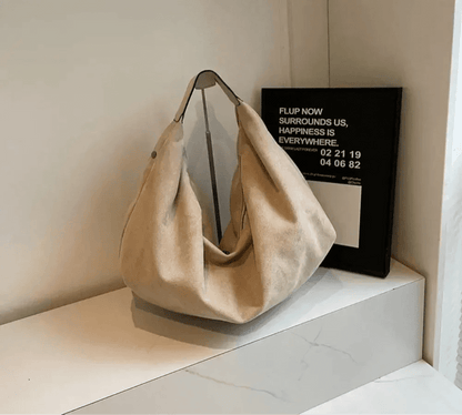 Beige suede hobo handbag with zipper and large capacity, displayed on a white shelf.